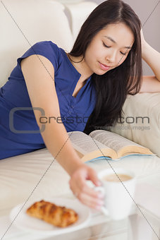 Pretty young asian woman lying on the sofa reading a book reaching for her coffee