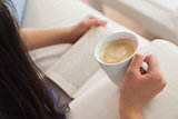 Woman lying on the sofa reading a book holding her coffee