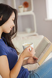 Young asian woman sitting on the sofa reading a book holding her coffee