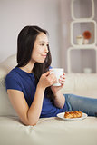 Young asian woman sitting on the sofa having coffee with a pastry