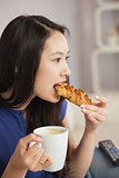 Young asian woman sitting on the sofa having coffee and eating a pastry