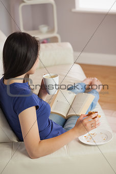 Young woman reading a book and eating pastry with coffee
