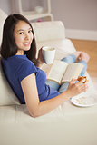 Young woman reading a book and eating pastry with coffee turning to the camera