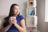 Thinking young asian woman sitting on the couch holding mug