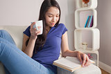 Reading young asian woman sitting on the couch holding mug