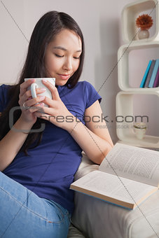 Reading asian woman sitting on the couch holding mug