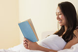 Happy young asian woman sitting in bed reading a book