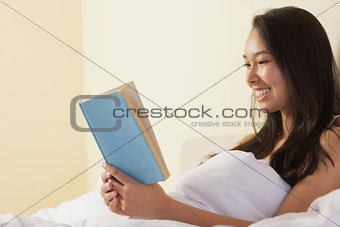 Happy young asian woman sitting in bed reading a book