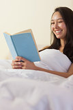 Pretty young asian woman sitting in bed reading a book