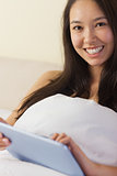 Pretty young asian woman sitting in bed using her digital tablet smiling at camera