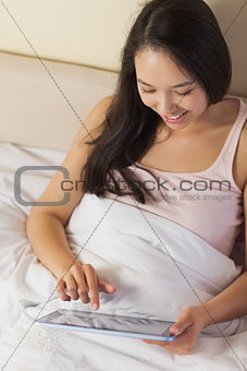 Cheerful young asian woman sitting in bed using her digital tablet