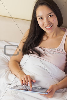 Cheerful young asian woman sitting in bed using her digital tablet looking up at camera
