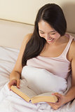 Cheerful young asian woman sitting in bed reading a book