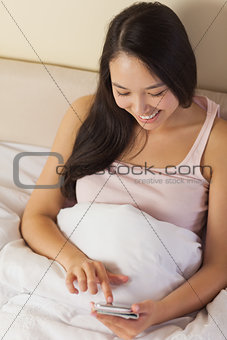 Cheerful young asian woman sitting in bed texting on her smartphone