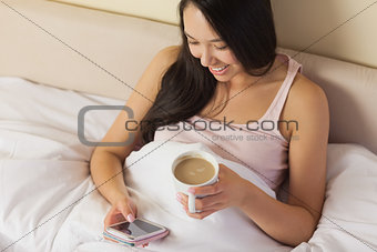 Cheerful young asian woman sitting in bed texting on her smartphone and drinking coffee