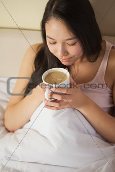 Cheerful young asian woman sitting in bed smelling her morning coffee