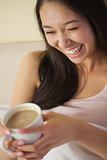 Laughing young asian woman sitting in bed holding her morning coffee