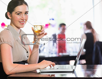 Happy businesswoman holding wine glass using laptop and looking at camera