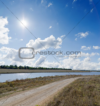 sun on cloudy sky over rural road and river near it