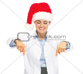Smiling doctor woman in santa hat pointing down on copy space