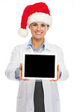 Smiling doctor woman in santa hat showing tablet pc blank screen