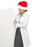 Happy doctor woman in santa hat looking on blank billboard