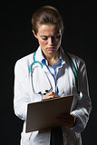 Serious doctor woman writing in clipboard on black background