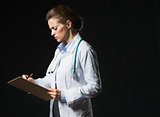 Serious doctor woman writing in clipboard isolated on black