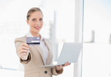 Smiling blonde businesswoman showing credit card holding laptop