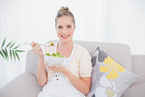 Smiling fresh model holding healthy salad sitting on sofa