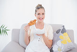 Smiling fresh model holding croissant sitting on sofa