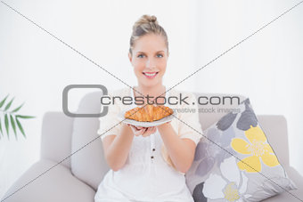 Cheerful fresh model holding croissant sitting on sofa