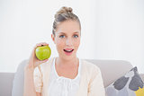 Surprised gorgeous model holding green apple sitting on sofa