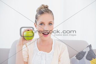 Surprised gorgeous model holding green apple sitting on sofa