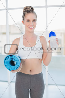 Smiling sporty blonde holding flask and exercise mat