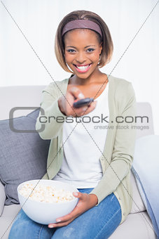 Cheerful cute woman sitting on sofa changing tv channel and holding popcorn