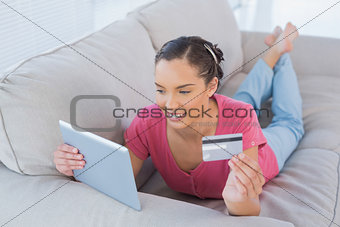 Attractive woman making an online payment with her credit card while lying on a sofa