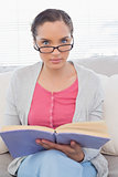 Serious woman with reading glasses sitting on sofa and holding a book