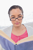 Focused woman with reading glasses sitting on sofa and reading a book
