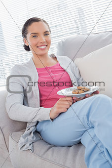 Cheerful woman sitting on sofa holding plate of cookies