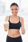 Delighted dark haired model in sportswear holding cup of coffee and giving thumb up