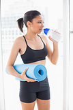 Serious dark haired model in sportswear carrying an exercise mat and drinking water