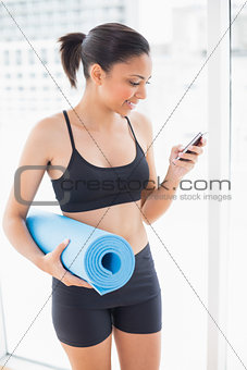Pleased dark haired model in sportswear carrying an exercise mat and using a mobile phone