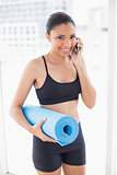 Fit dark haired model in sportswear carrying exercise mat and making a phone call