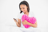 Pleased dark haired model cuddling a pillow and using a mobile phone