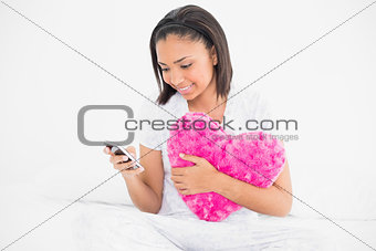Pleased dark haired model cuddling a pillow and using a mobile phone