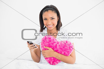 Delighted young dark haired model holding a pillow and a mobile phone