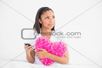 Thoughtful young dark haired model holding a pillow and a mobile phone