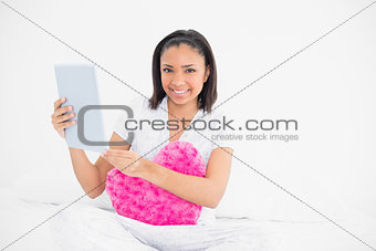 Smiling young dark haired model cuddling a pillow and using a tablet pc