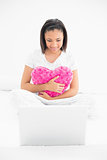 Concentrated young dark haired model cuddling a pillow and looking at a laptop